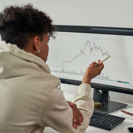 Back view of focused young male trader looking at computer screen, studying trading charts while working from homeの写真素材