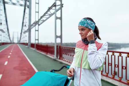 Young woman using earbuds while standing on the bridge with a baby carriage, ready for a jog on a...