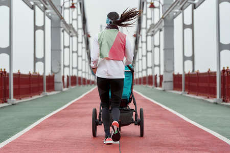 Young mother sportswoman jogging with a baby carriage on a bridge in the city on a cloudy dayの写真素材