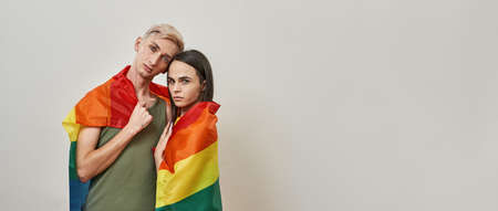 Couple of young transgenders wrapped in colorful rainbow flag of LGBT looking at camera while posing together isolated over light backgroundの写真素材