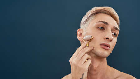 Portrait of androgynous transgender young man with light hair using quartz roller for face massage and looking away while posing isolated over dark blue backgroundの写真素材