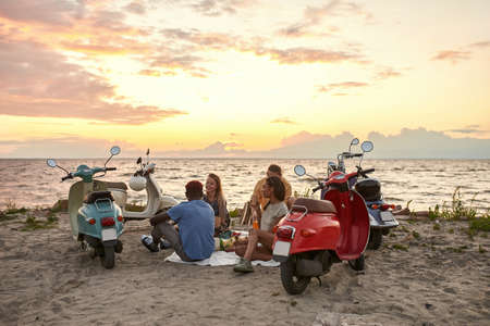 Young caucasian people, motorcycle riders enjoying the view while having picnic on the beach at sunsetの写真素材