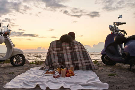 Rear view of loving couple enjoying the view, sitting together covered with blanket on the beach outside the cityの写真素材