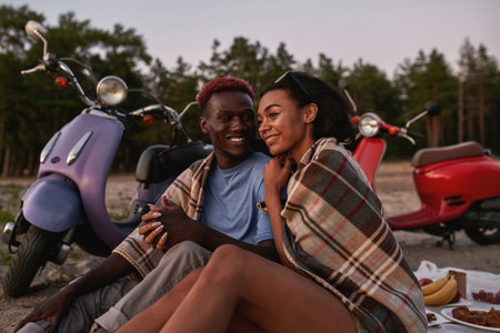 Lovely young diverse couple smiling, relaxing together on the beach covered with blanket in the eveningの写真素材