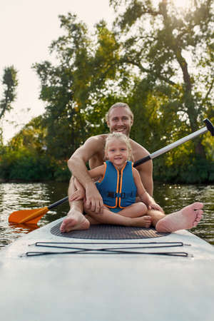 Portrait of father together with his little daughter smiling at camera, relaxing, sup surfing on a river on a summer dayの写真素材