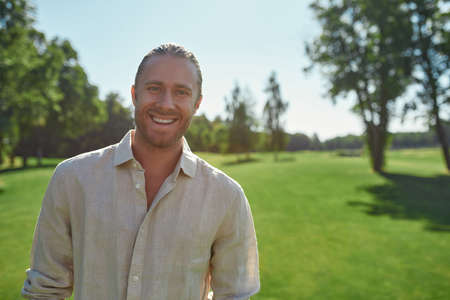 Portrait of cheerful young man smiling at camera while standing in the park on a summer day