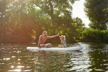 Father giving high five to his little daughter while relaxing, sup surfing on a river surrounded by...
