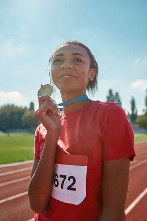 Portrait of young female athlete champion smiling away and holding her gold medal while standing on race trackの写真素材