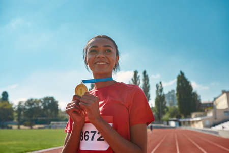 Joyful young female athlete smiling away and holding her gold medal while standing on race trackの写真素材