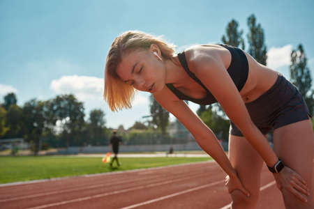 Exhausted sporty young woman taking a break while training, running on the track at stadium on a...