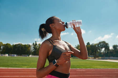Professional young female runner drinking water after training, standing on the track at stadium on a sunny dayの写真素材