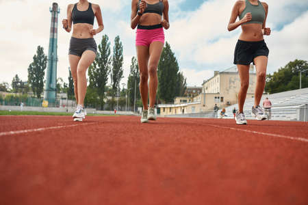 Cropped shot of three professional young female runners in sportswear running together on track field at the stadiumの写真素材