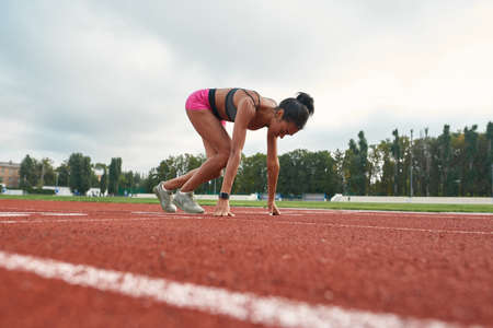 Side view of professional young female runner in sportswear starting race on track field at stadiumの写真素材