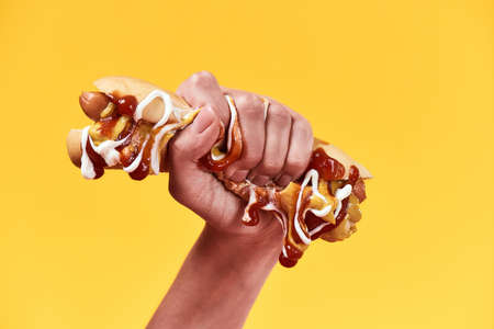 Female hand squeezing out junk food, hot dog with mustard and ketchup isolated over yellow backgroundの写真素材