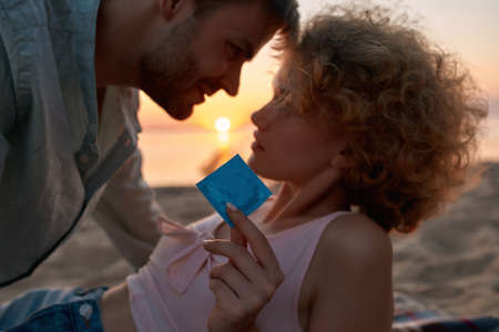 Portrait of young couple making love on the beach at sunset. Woman is holding and showing a condom...