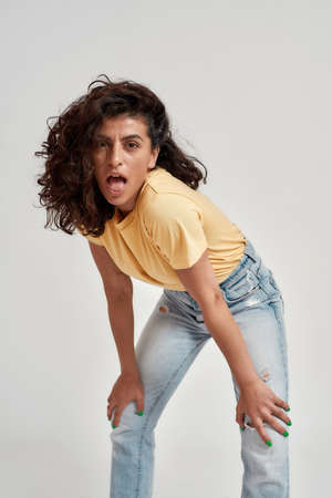 Playful young female model with dark curly hair in casual wear looking at camera, sticking out tongue while posing isolated over gray backgroundの写真素材