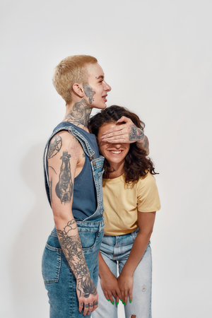 Two diverse young women, lesbian couple posing together isolated over gray background. Woman holding hand over her girlfriend eyesの写真素材