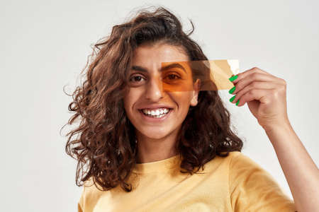 Happy woman with curly hair holding orange film in front of her eyes, posing isolated over light backgroundの写真素材