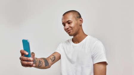 Portrait of tattooed mixed race young man in t shirt taking selfie using smartphone while standing...