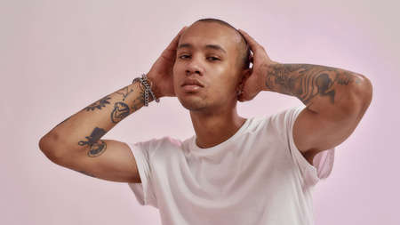 Portrait of confident young mixed race tattooed guy in white t shirt holding hands on his head while posing for camera isolated over pink backgroundの写真素材