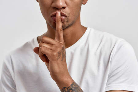 Cropped shot of dark skinned young man showing shh gesture, keeping a secret, posing isolated over white backgroundの写真素材