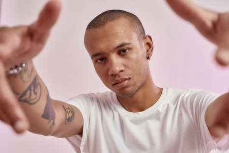 Portrait of cool young mixed race tattooed guy in white t shirt looking at camera, reaching out his arms while posing isolated over pink backgroundの写真素材