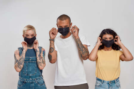 Portrait of three diverse young people in casual wear putting medical masks on face, posing together isolated over gray backgroundの写真素材