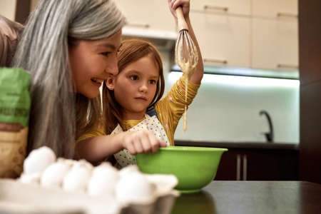 Girl looking on whisk with dough with grandmother