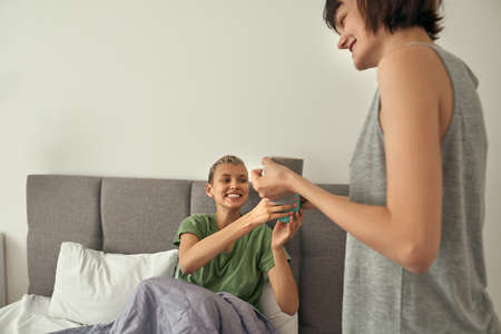 Girl giving cup with coffee to girlfriend in bed