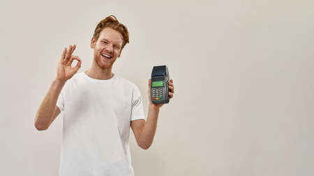 Young redhead man holds payment terminal in front of him, shows ok with his handの写真素材