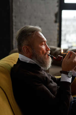 Senior man light tobacco pipe with match on sofa