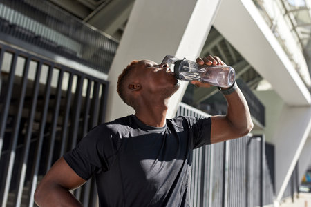 Tired black sportsman drinking water from bottleの写真素材