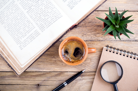 Coffee cup, open book, succulent aloe, pen, notebook and magnifier on rustic wooden background. Office desk table. Educations concept. Top view.の写真素材