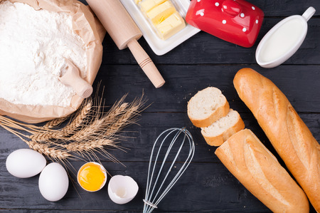Baking background. Flour in paper bag, eggs, butter, milk, rolling pin and whisk on dark wooden...