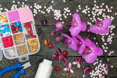 Box with beads, spool of thread, plier and glass hearts to create hand made jewelry on old wooden background. Handmade accessories. Orchid flowers. Top viewの写真素材