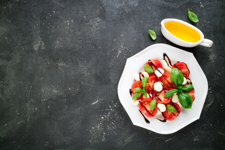 Caprese salad. Mozzarella cheese, tomatoes, olive oil, basil herb leaves on dark background. Italian food. Top viewの写真素材