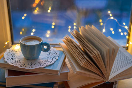 Coffee cup, open book, fairy lights on window sill on blue blurred background. Concept warm and cozy home, reading books, hygge, evening rest and comfort leisureの写真素材