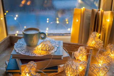 Coffee cup, open book, sparkle lighting garland on window sill on blue blurred background. Concept warm and cozy home, reading books, hygge, evening rest and comfort leisureの写真素材