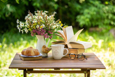 Bouquet of meadow flowers, croissant, cup of tea or coffee, books on table in summer idyllic garden. Rest in garden, reading books, breakfast, vacations in nature concept. Summertime in garden on backyardの写真素材