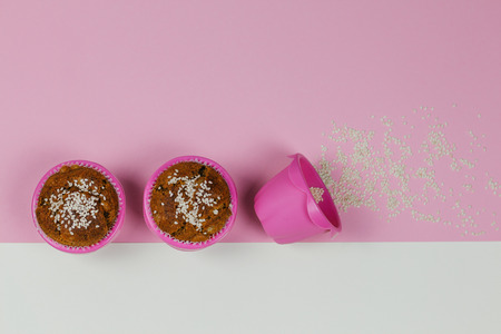 Two banana muffins sprinkled with sesame in pink baking cups and one baking cup sideways with the trail of sesame on pink and white background.の写真素材