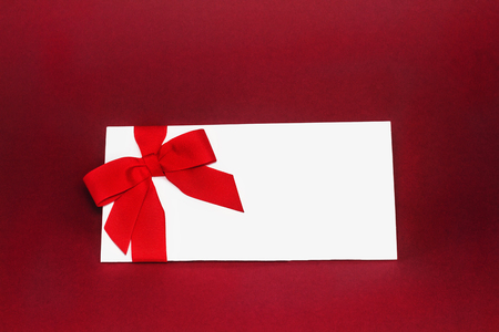 Isolated white gift card with silk red ribbon bow on deep red background. Cut out.の写真素材