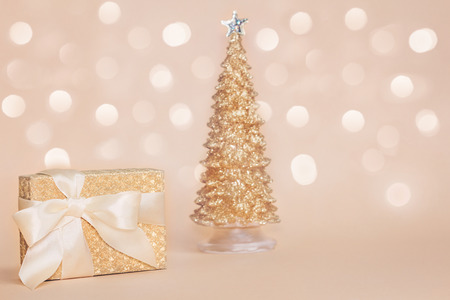 Beautiful golden present box with a silk bow against blurred gold sparkling christmas pine tree on pastel background with beautiful shining bokeh. Copy space.の写真素材