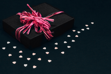 Vintage black gift box with pink ribbon bow on black background sprinkled with heart shaped confetti. Copy space.