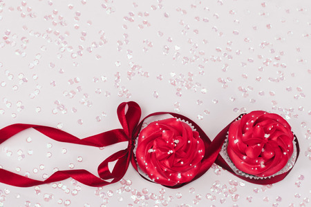 Two cupcakes with red icing decorated with red ribbon with a bow on white background with sprinkles. Womens day concept. Copy space.の写真素材