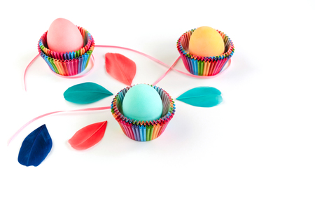 Bright creative Easter decoration of colorful eggs in raibow colored paper forms for cupcakes on white background decorated with feathers. Copy space.の写真素材