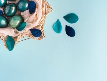 Easter background with eggs decorated in blue, turquoise and gold in nest with colorful blue and turquoise feathers. Top view. Copy space.の写真素材