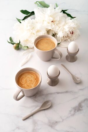 Boiled eggs in ceramic egg cups, two cups of coffee with ceramic spoons on background of beautiful white peony. Breakfast concept. Copy space.の写真素材