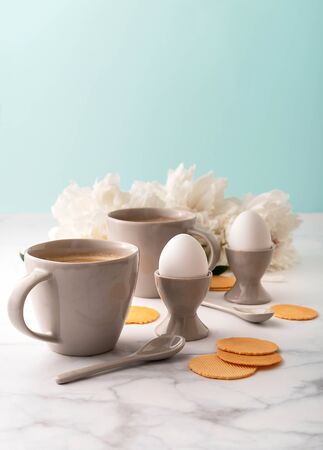 Boiled eggs in ceramic egg cups, two cups of coffee with ceramic spoons on background of beautiful white peony. Breakfast concept. Copy space.の写真素材