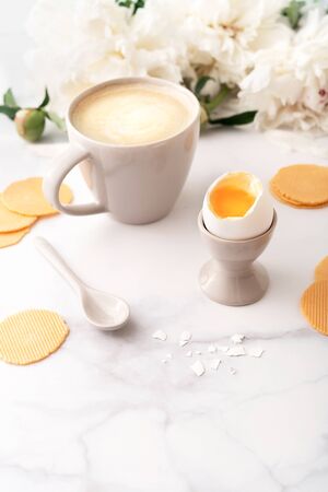 Soft-boiled egg with liquide orange yolk in ceramic egg cup, cup of coffee with ceramic spoon and thin crispy corn chips on background of beautiful white peony. Breakfast concept. Copy space.の写真素材
