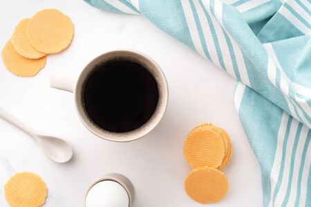Top view of boiled egg in ceramic egg cup, cup of coffee and thin crispy corn chips on background of beautiful white peony. Breakfast concept. Copy space.の写真素材